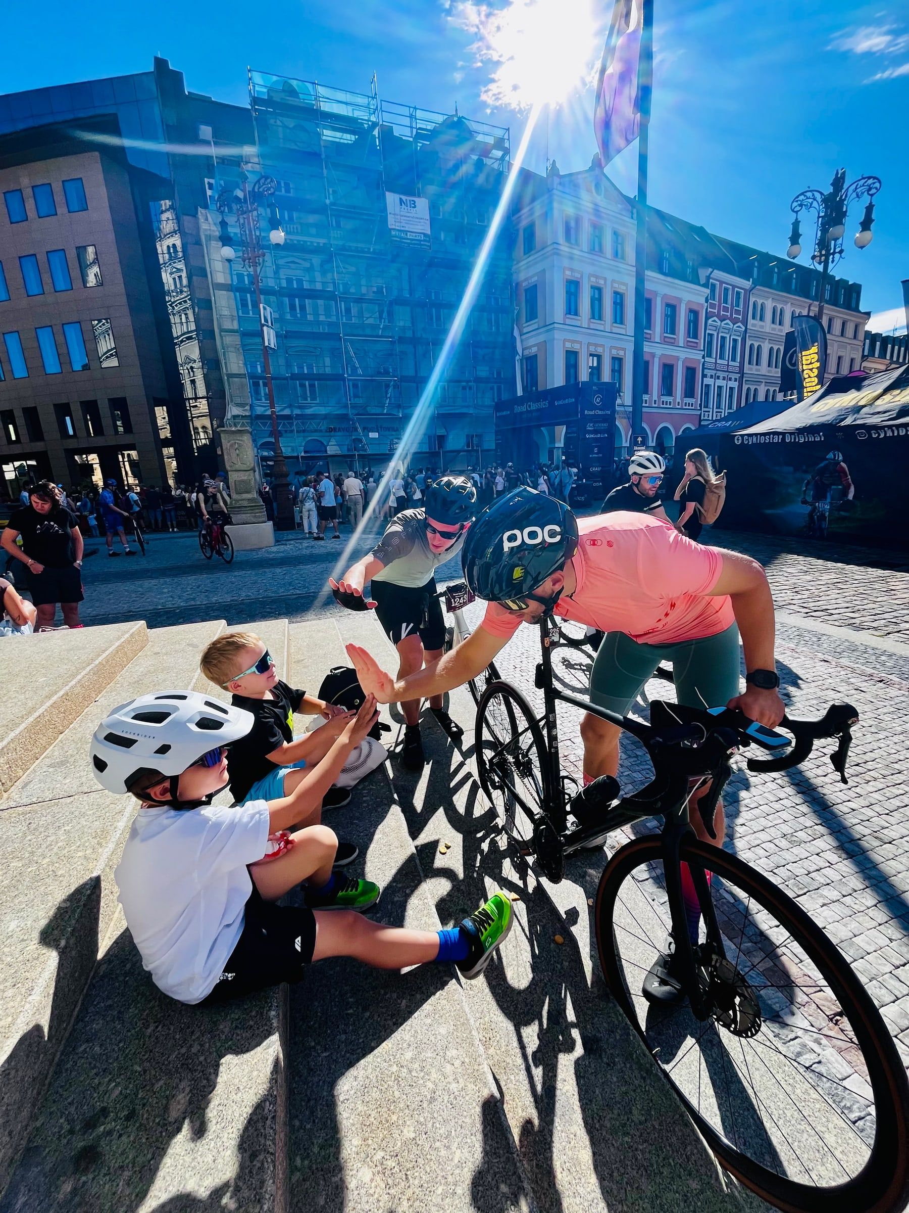 Cyklista v růžovém dresu si na náměstí při sportovní akci plácá s dětmi v helmách sedícími na schodech.